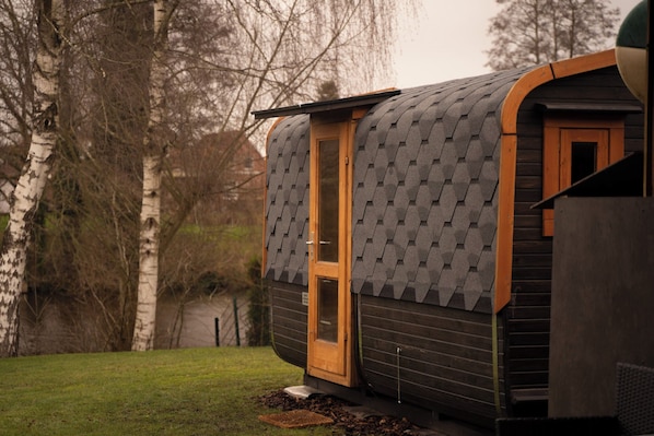 A small, modern cabin with a curved roof and a wooden door, surrounded by trees and a grassy area.