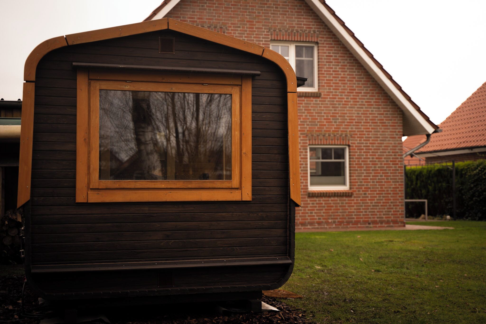A small wooden cabin with a window and a door, situated in front of a brick house.