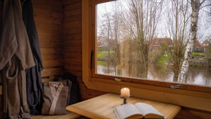 Una stanza in una baita di legno con una panca, un tavolo con un libro aperto e una finestra che si affaccia su un corpo d'acqua e delle case.