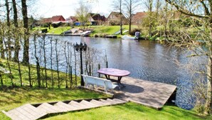 Una terrazza sul fiume con tavolo e panche, una barca ormeggiata nell'acqua e case sullo sfondo.