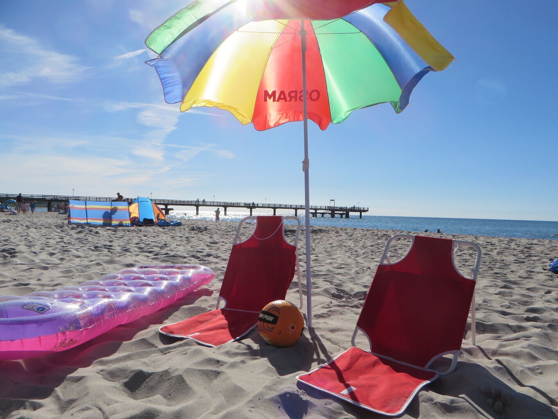 Ein Strand mit roten Liegestühlen, einem bunten Sonnenschirm und einem pinken Luftmatratze.