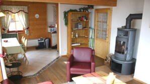 A cozy living room with a fireplace, a red armchair, a wooden bookshelf, and a dining area with a table and chairs. - Exclusive vacation home in the Nockberge with a sauna and fireplace (Patergassen)