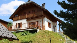 A two-story wooden house with a balcony, a staircase leading up to the entrance, and a view of the surrounding greenery. - Exclusive vacation home in the Nockberge with a sauna and fireplace (Patergassen)