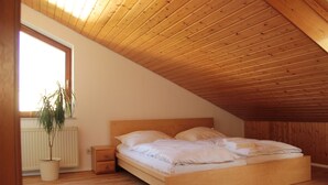Un dormitorio con techo de madera inclinado, una cama individual con ropa de cama blanca, una mesita de noche, una planta en maceta y una ventana que deja entrar luz natural.