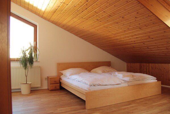 A bedroom with a sloped wooden ceiling, a single bed with white bedding, a nightstand, a potted plant, and a window providing natural light.