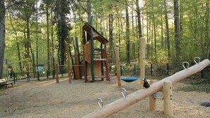 Un parque infantil con un sube y baja, estructuras para escalar y columpios en un área boscosa.