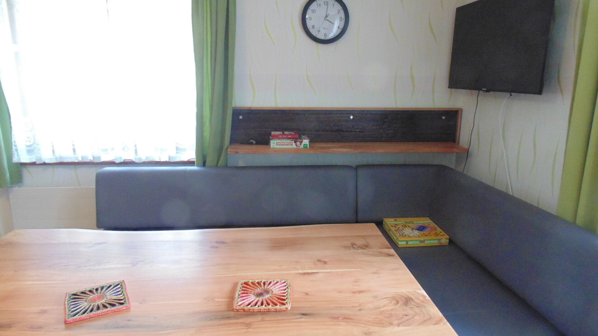 A room with a wooden table, two decorative items, a grey sofa, a shelf with Monopoly boxes, and a clock on the wall.