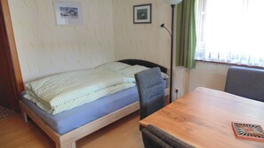 A small, simple room with a single bed, a chair, a table, and a lamp.