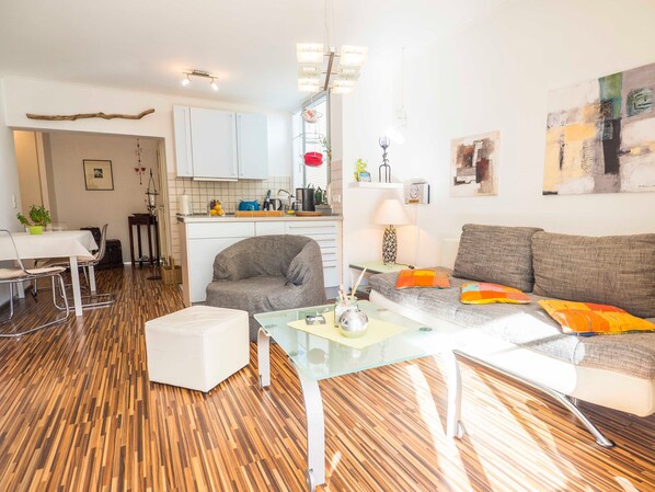 A modern living room with a grey sofa, a glass coffee table, and a white armchair. There is a kitchen area with white cabinets and a dining table with chairs.