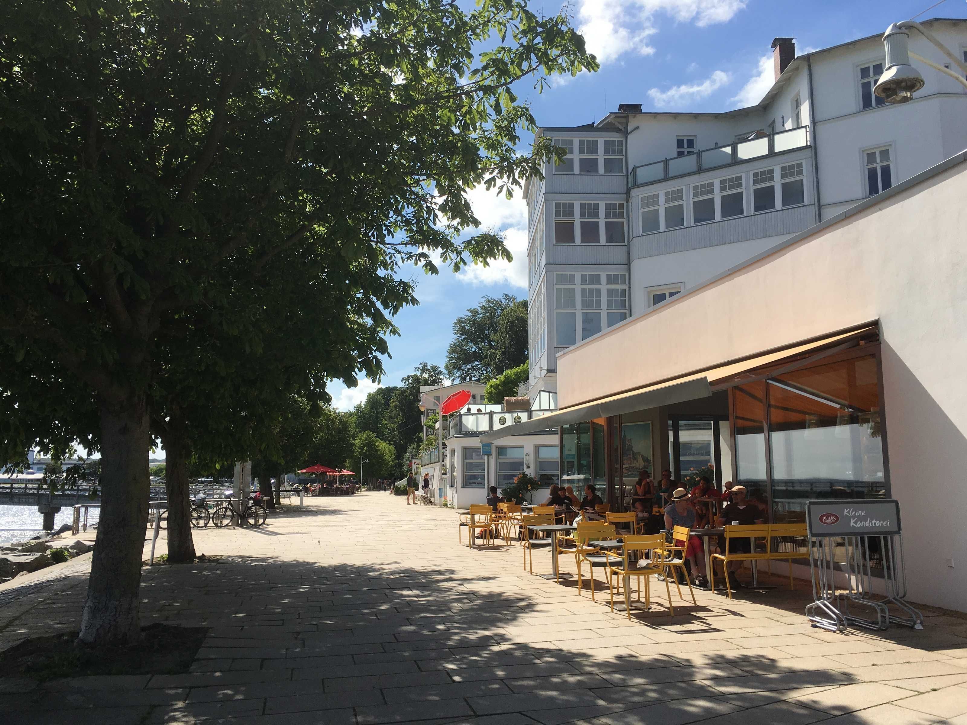 An outdoor café with yellow chairs and tables, a view of the water, and a building with large windows in the background.