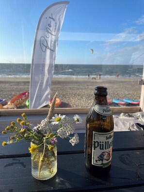 A bottle of Pülled pulle and a vase of flowers on a table with a view of the beach and ocean.