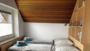 Ein kleiner, gemütlicher Raum mit schrägem Dach und Fenster. Es gibt ein hölzernes Bett mit zwei Kissen und einer Decke, einen Holzschrank und einen Schreibtisch mit einer Lampe.