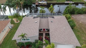 A two-story house with a tiled roof, a front porch, and a small garden with red flowers and palm trees.