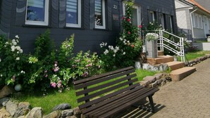 Une maison à deux étages, à l’extérieur sombre, dotée de nombreuses fenêtres et d’un jardin devant avec des fleurs et un banc.