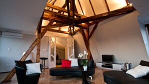 A modern loft-style living room with exposed wooden beams, a curved black sofa, a black armchair, and a small side table with a potted plant.
