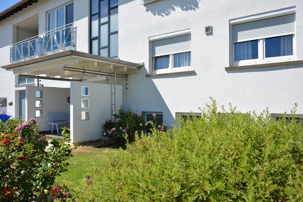 A modern two-story building with a balcony, a covered entrance, and a garden area with flowers.