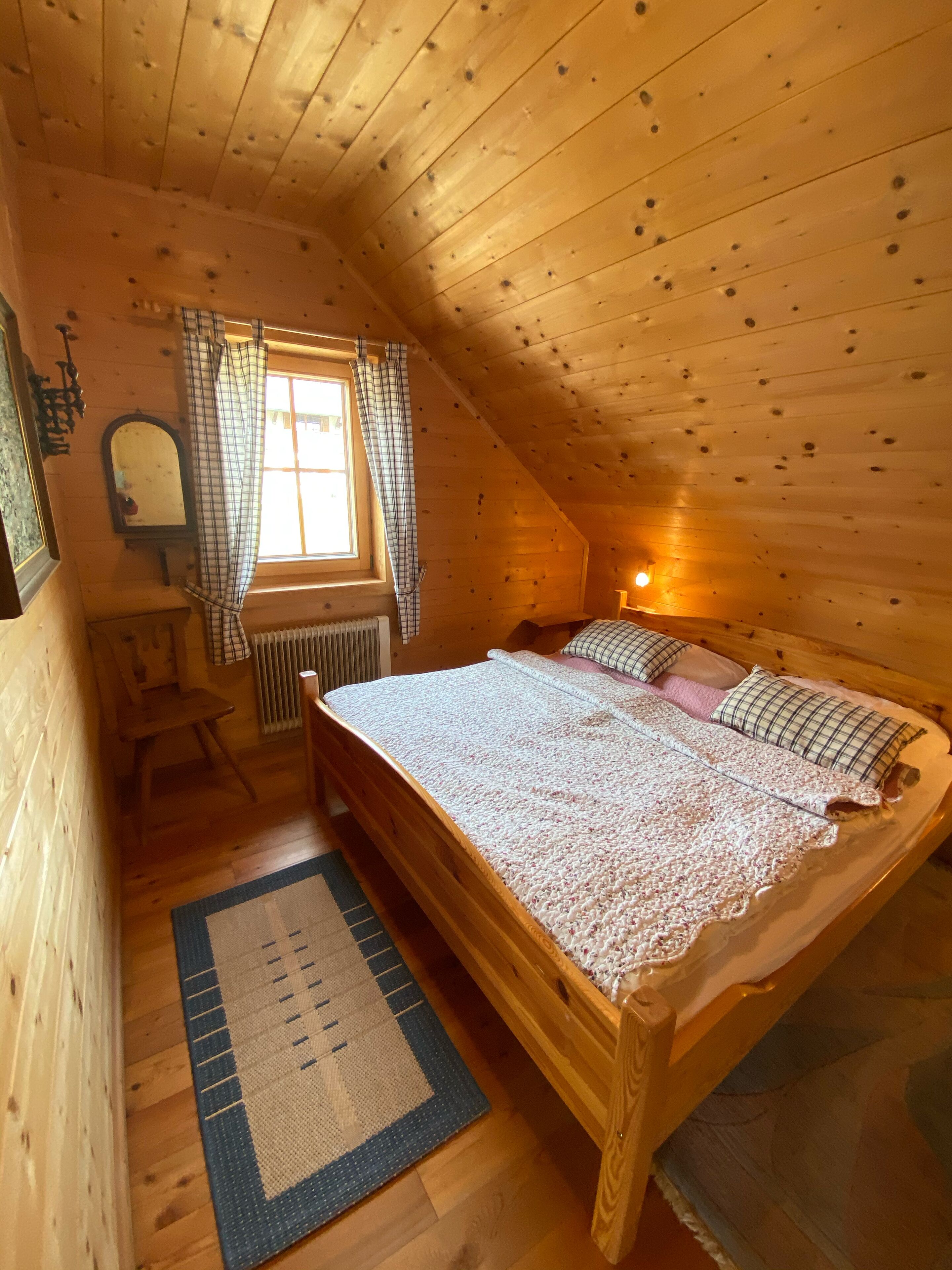 Ein gemütliches Schlafzimmer im Dachgeschoss mit schrägen Holzwänden, einem großen Bett mit gemustertem Überwurf, einem Fenster mit Vorhängen, einer Heizung und einem kleinen Stuhl.