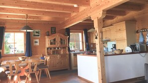Interior de una cabaña de madera con comedor, cocina y ventanas amplias.