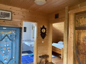 A wooden interior with a bathroom visible through an open door, a bed with a blue and white checkered blanket, and a small stool.