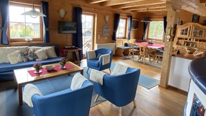 Interior acogedor de una cabaña con techo de madera, un sofá, sillas azules, una mesa de comedor y una zona de cocina.