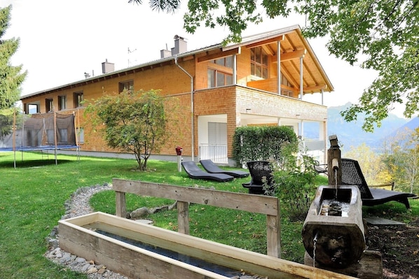 Ein modernes Haus mit einer Holzterrasse, einem Trampolin und einem kleinen Garten.