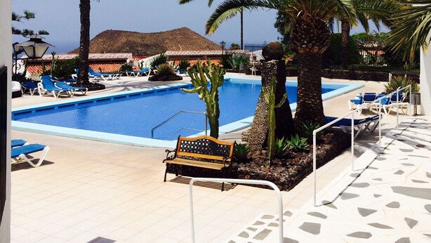 A pool area with lounge chairs and a bench, surrounded by palm trees. - Beautiful apartment 98 sqm with sea view 2 bedrooms, balcony + terrace, pool, free wifi (Chayofa)
