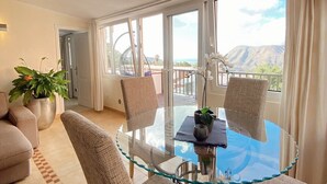 A modern living room with a glass dining table, two chairs, a sofa, and a large window offering a view of the mountains. - Beautiful apartment 98 sqm with sea view 2 bedrooms, balcony + terrace, pool, free wifi (Chayofa)