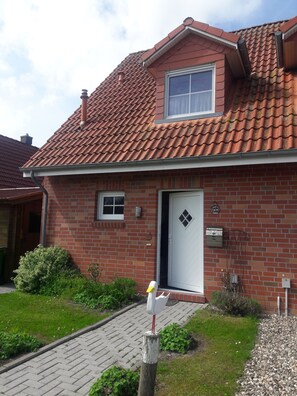 Une maison à deux étages, construite en briques, avec un toit recouvert de tuiles rouges, une porte d’entrée dotée d’une petite fenêtre et une boîte aux lettres située sur le côté droit de la porte.