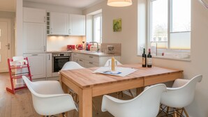Una cocina moderna con armarios blancos, una mesa de comedor de madera y sillas blancas. Hay una tetera roja, una cafetera y botellas de vino en la encimera.