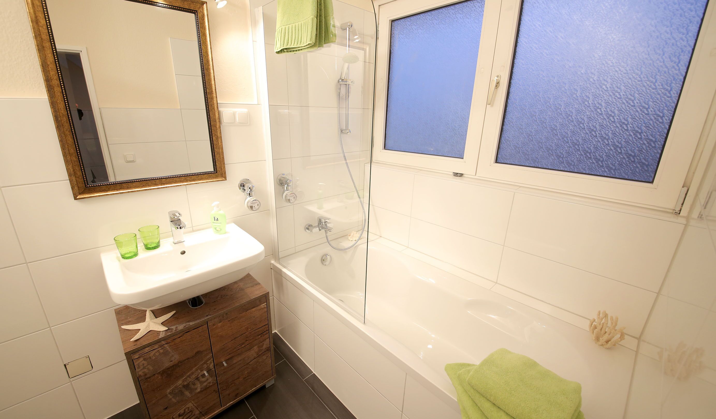 Ein Badezimmer mit einem weißen Waschbecken, einem Spiegel und einer Dusche. Ein grünes Handtuch und eine Meerstern-Dekoration liegen auf dem Waschtisch.