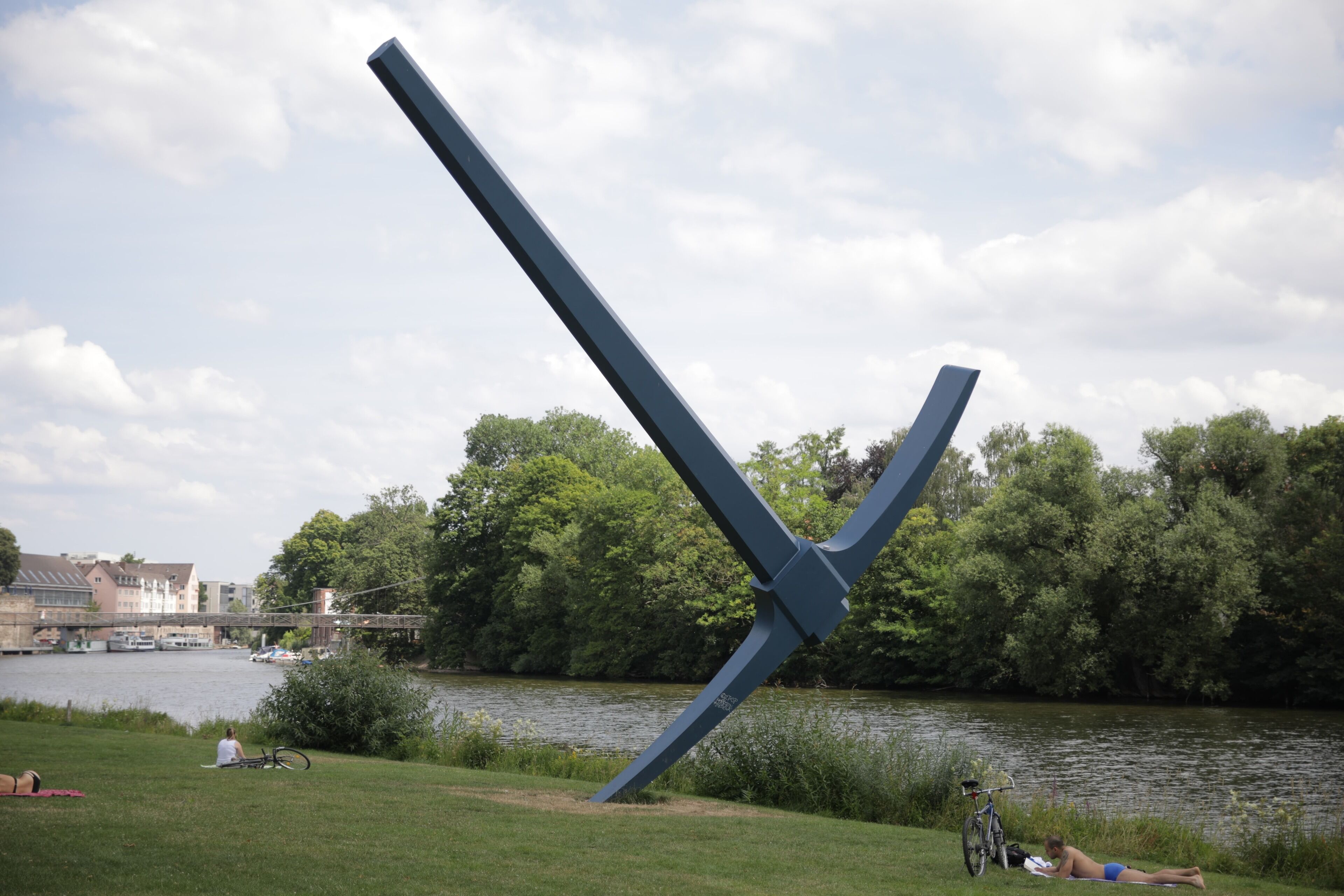 En park med en stor skulptur av metall, en flod och en bro i bakgrunden.