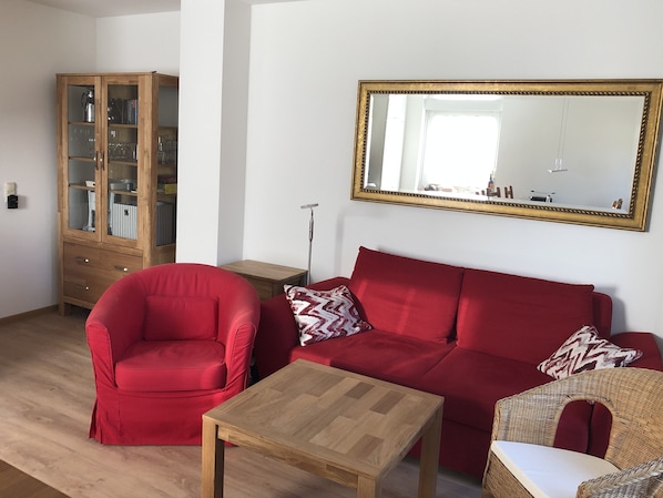 Una sala de estar con un sofá rojo, un sillón rojo, una mesa de centro de madera, una silla de mimbre y un espejo grande en la pared.