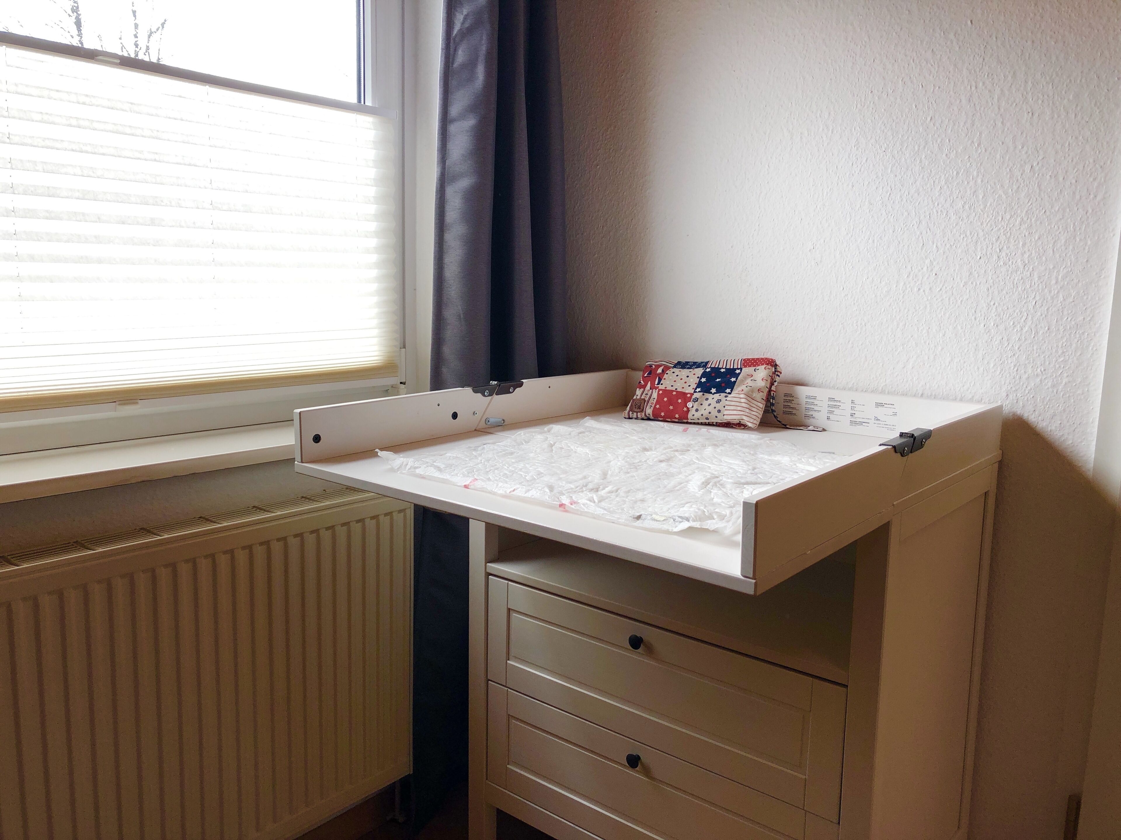 Ein Wickeltisch für Kinder mit einer gesteppten Auflage, ein Heizkörper und ein Fenster mit Jalousien.