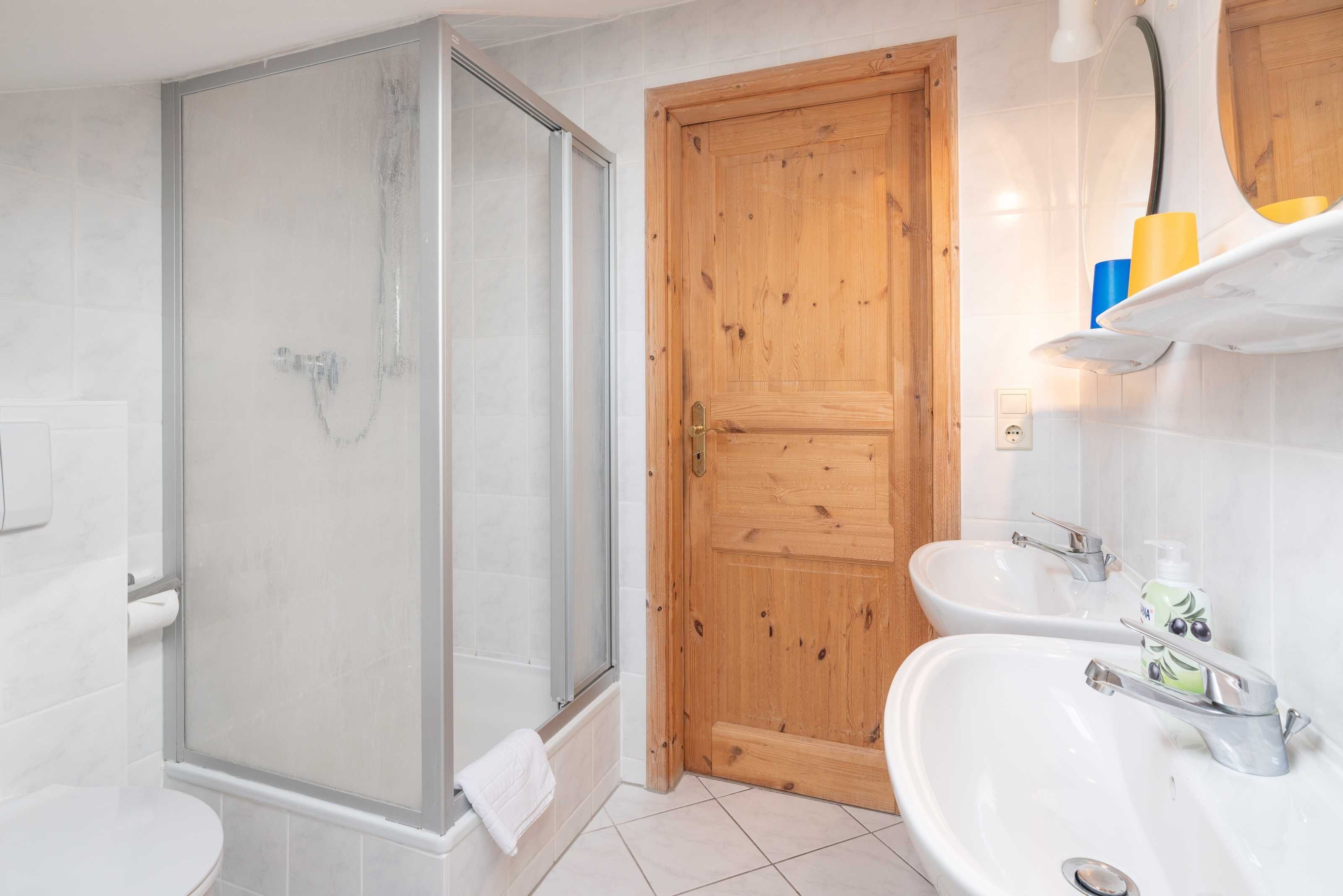 A bathroom with a glass-enclosed shower, a wooden door, and two sinks with faucets.