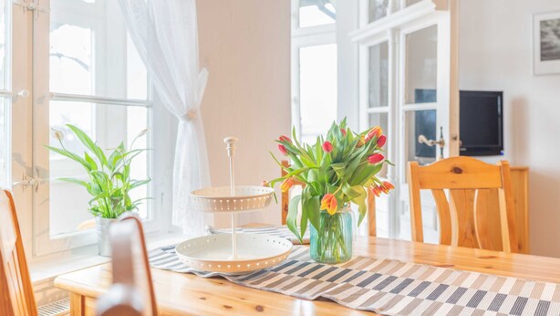 A dining area with a wooden table, two chairs, a vase with flowers, a tray stand, and a potted plant.