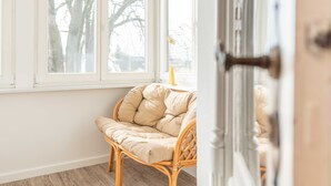 A room with a wicker chair by a window, a door with a brass handle, and a wooden floor.