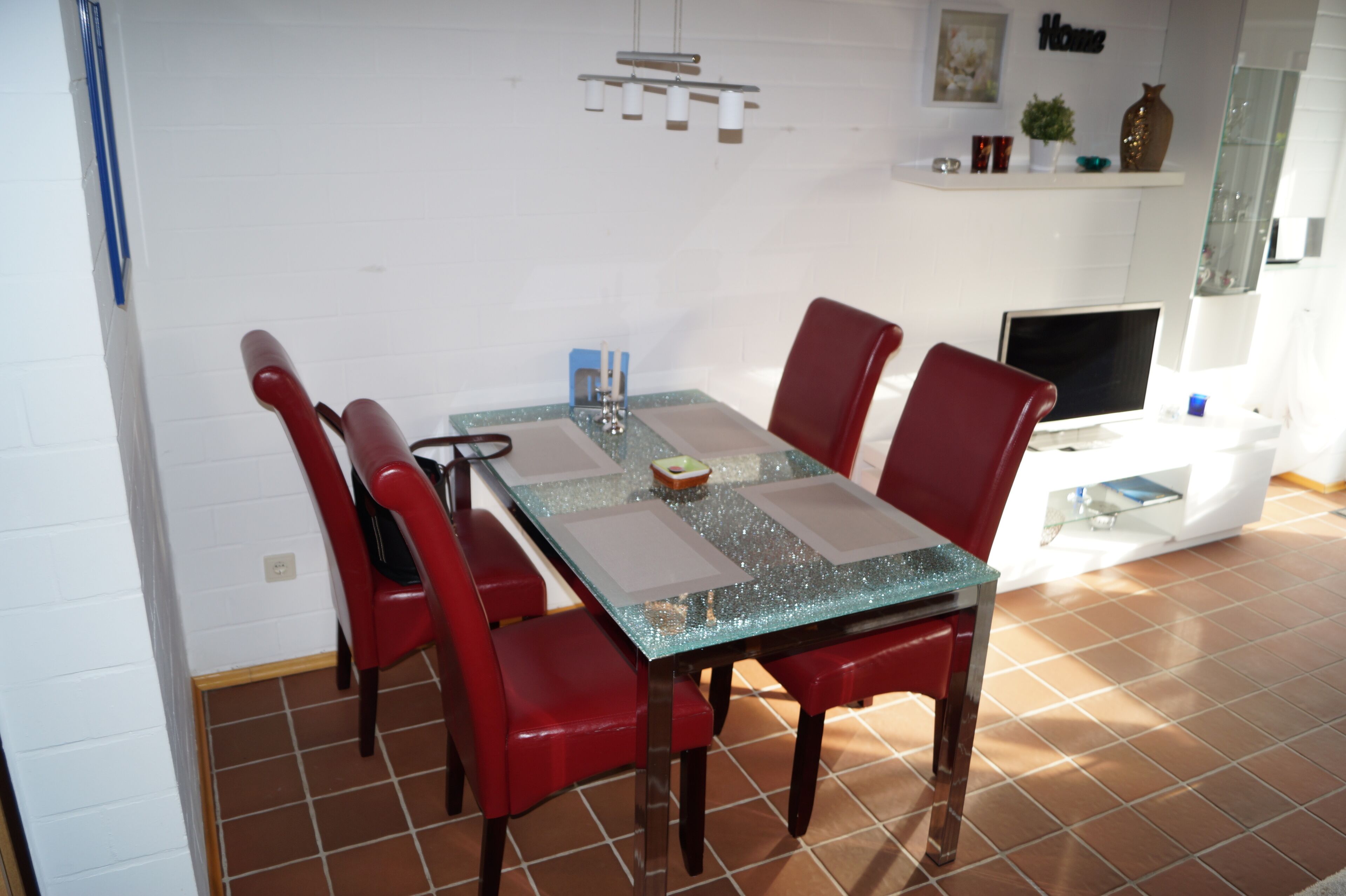 A dining area with a glass-top table, red chairs, a white wall, and a shelf with decorative items.