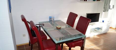 A dining area with a glass-top table, red chairs, a white wall, and a shelf with decorative items.