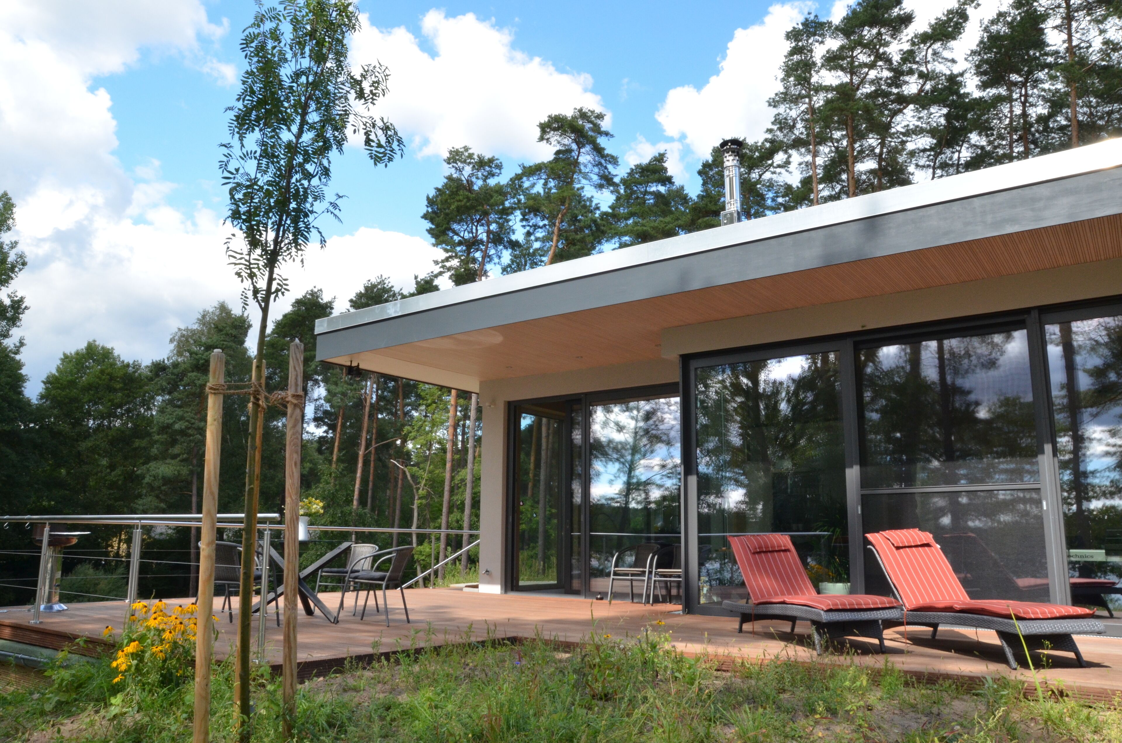 A modern house with a glass facade, outdoor seating area, and a deck overlooking a forested area.