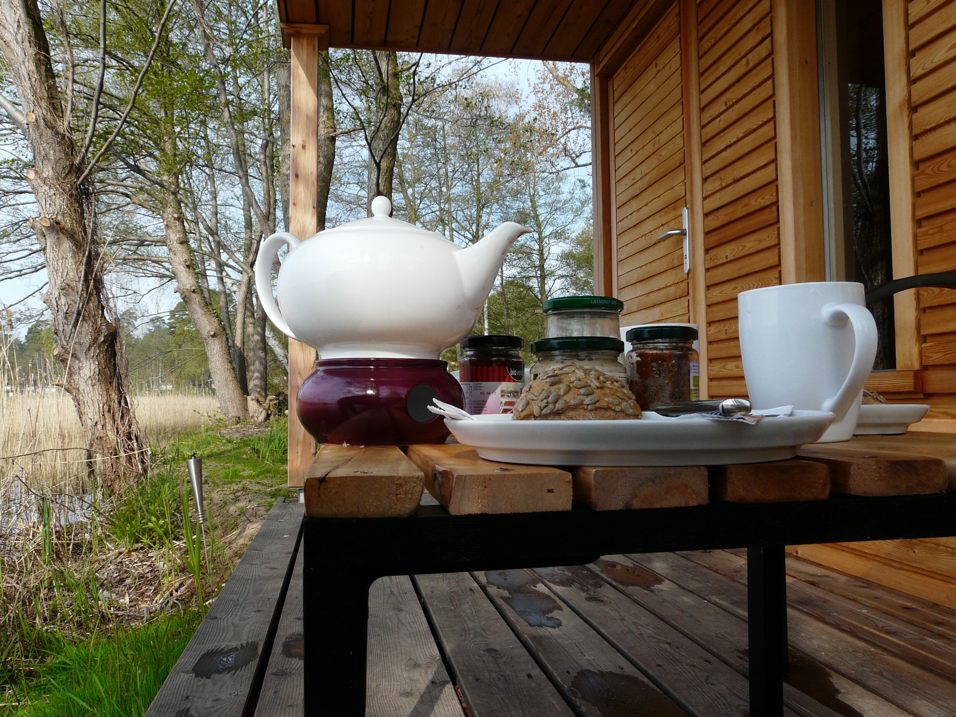 A wooden table with a white teapot, a white cup, and a plate with cookies and jars of jam on a wooden deck.