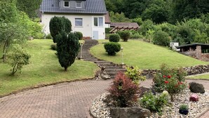 Una casa amb teulada inclinada, un jardí davanter amb un camí que condueix a l'entrada i una petita zona de jardí amb plantes diverses i una paret de pedra.