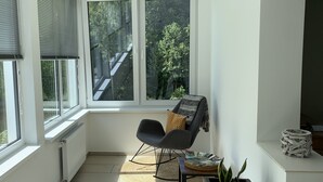 Habitació lluminosa amb grans finestres, una cadira negra, una taula petita amb revistes i una cistella de vimet.