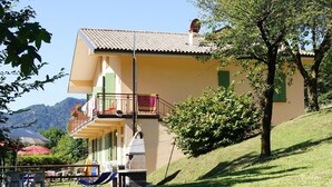 Un bâtiment à deux étages doté d’un balcon, d’un banc en pierre et, au fond, d’une colline verdoyante.