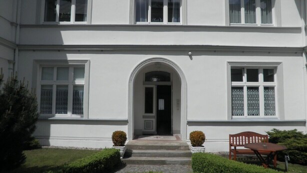 A white building with a central entrance, two potted plants on either side, and a bench in front. - Exclusive apartment (3 rooms, 85 sqm) in a quiet location, only 50m to the beach! (Heringsdorf)