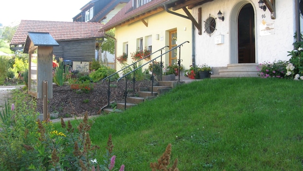 Ein traditionelles Haus mit gelber Fassade, einer hölzernen Treppe, die zu einer Tür führt, und einem gepflegten Garten mit verschiedenen Pflanzen und Blumen.