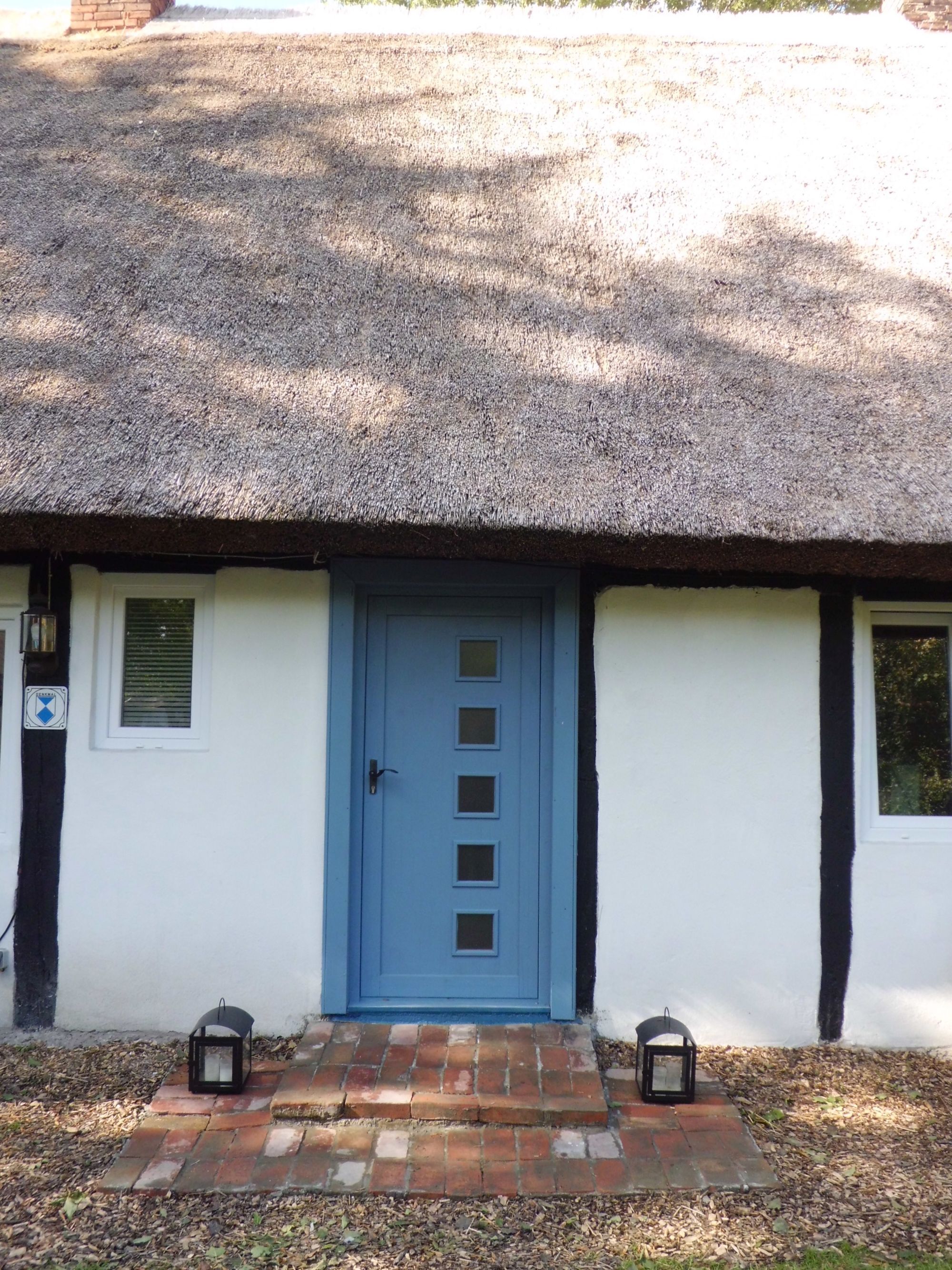 A thatched-roof cottage with a blue door, a small window with shutters, and two lanterns on the brick steps.