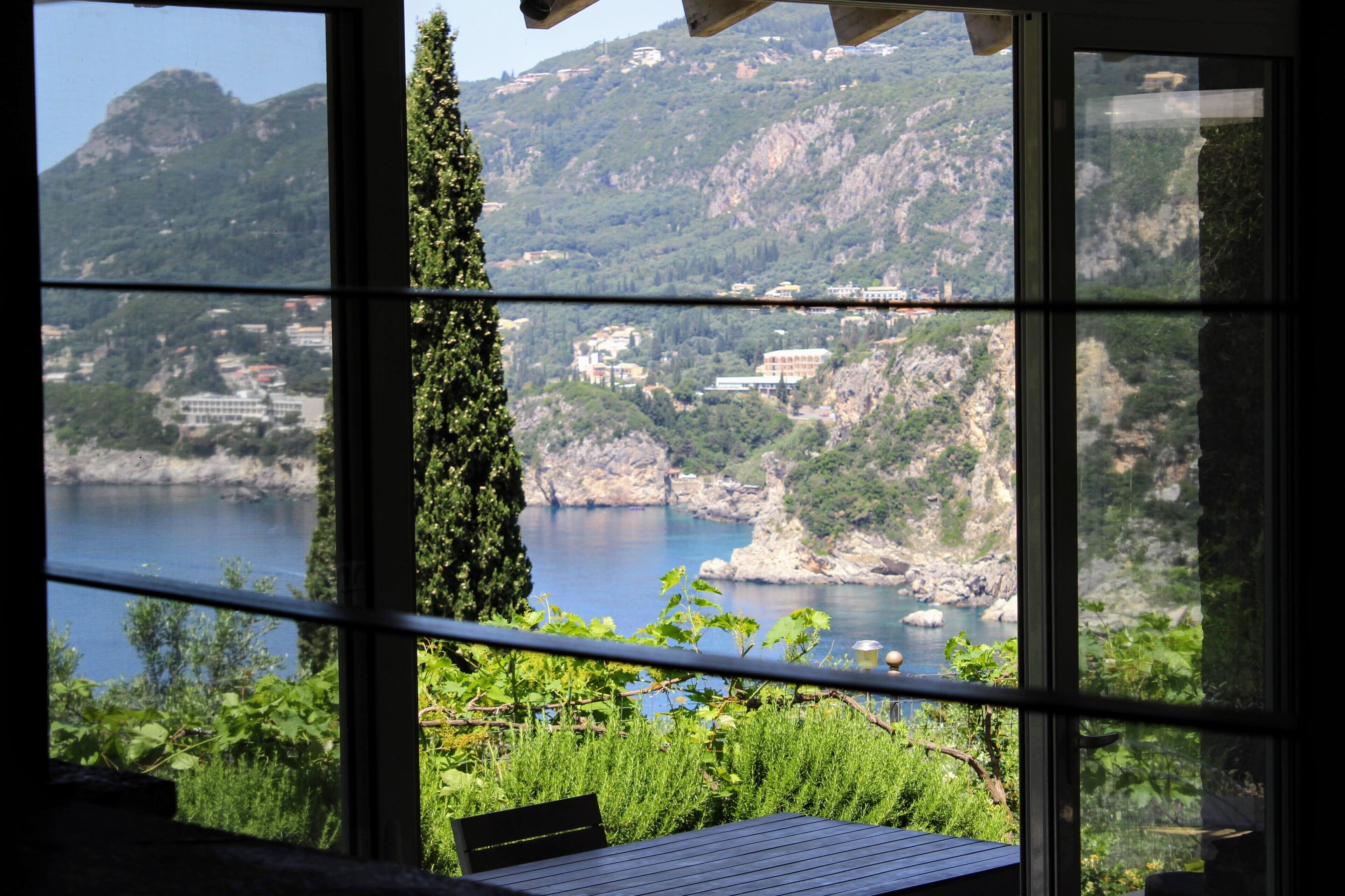 A view through a window with a table and chairs, overlooking a body of water and a mountainous landscape with buildings.