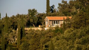 A stone building with a tiled roof, surrounded by dense trees and a tall cypress tree.