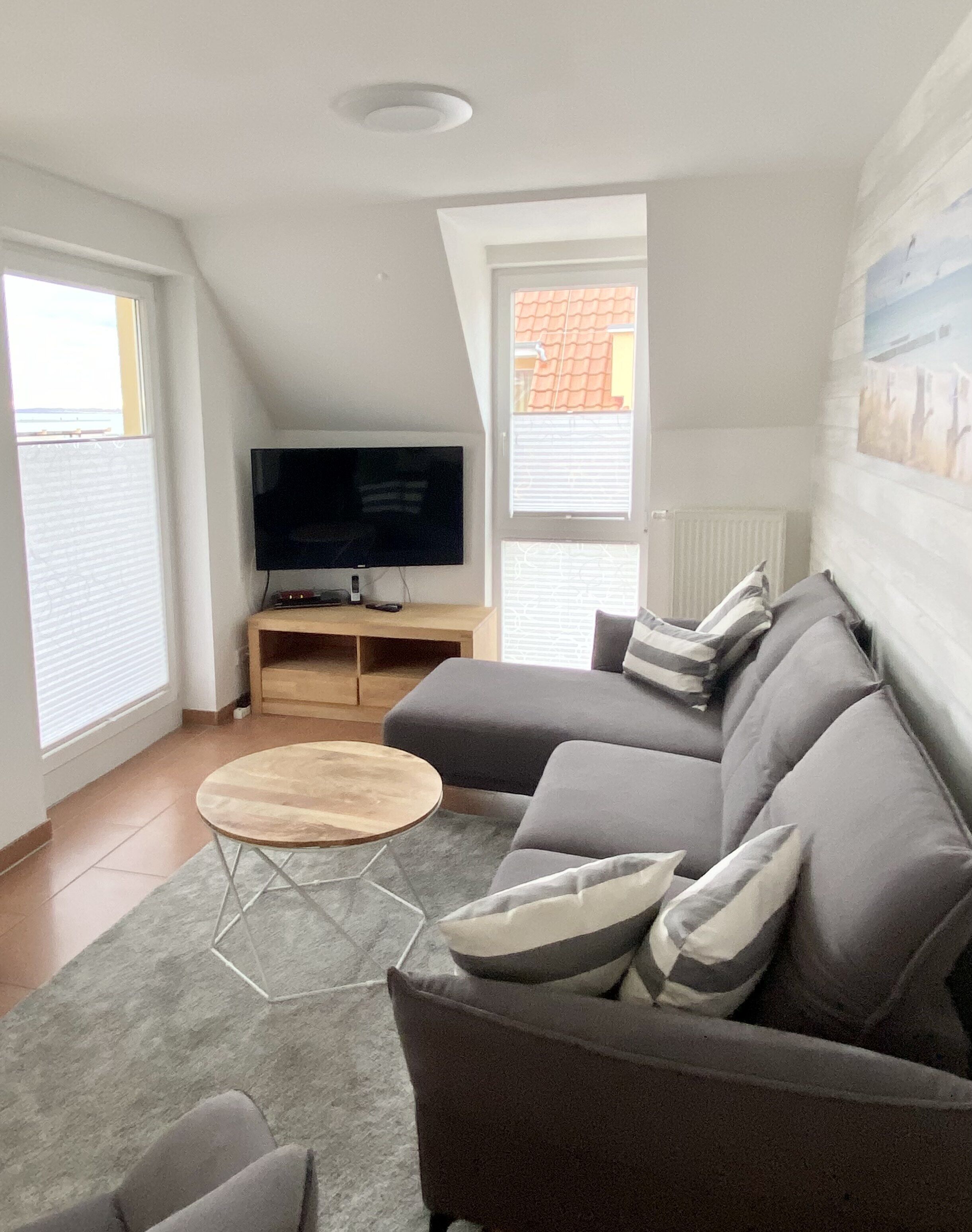 Ein modernes Wohnzimmer mit einem grauen Ecksofa, einem Holztisch, einem Flachbildfernseher auf einem Holzständer und einem grauen Teppich.