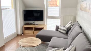 Ein modernes Wohnzimmer mit einem grauen Ecksofa, einem Holztisch, einem Flachbildfernseher auf einem Holzständer und einem grauen Teppich.